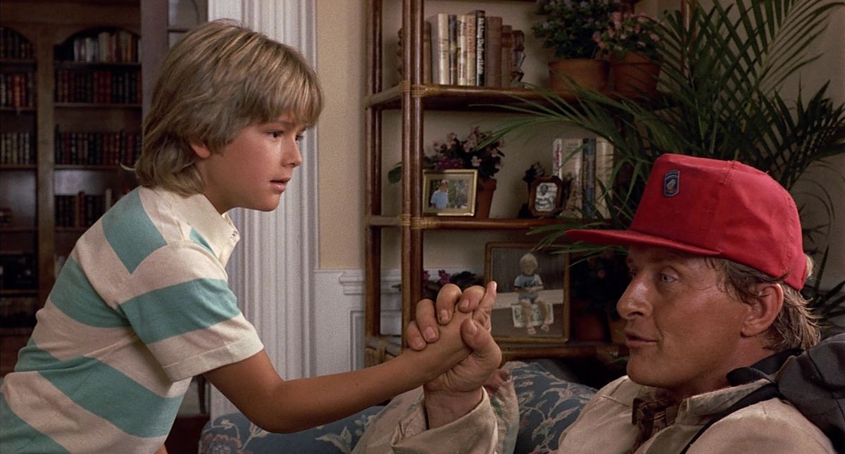 Rutger Hauer sits in a chair and holds a child’s hand in Blind Fury while looking away. He wears a flat-billed red hat.