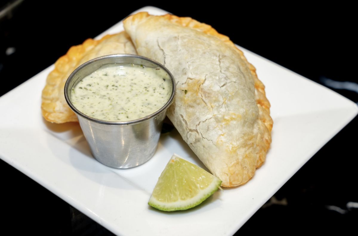 This photo shows a beef empanada beside a small container of chimichurri aioli sauce. A lime wedge decorates the plate. 
