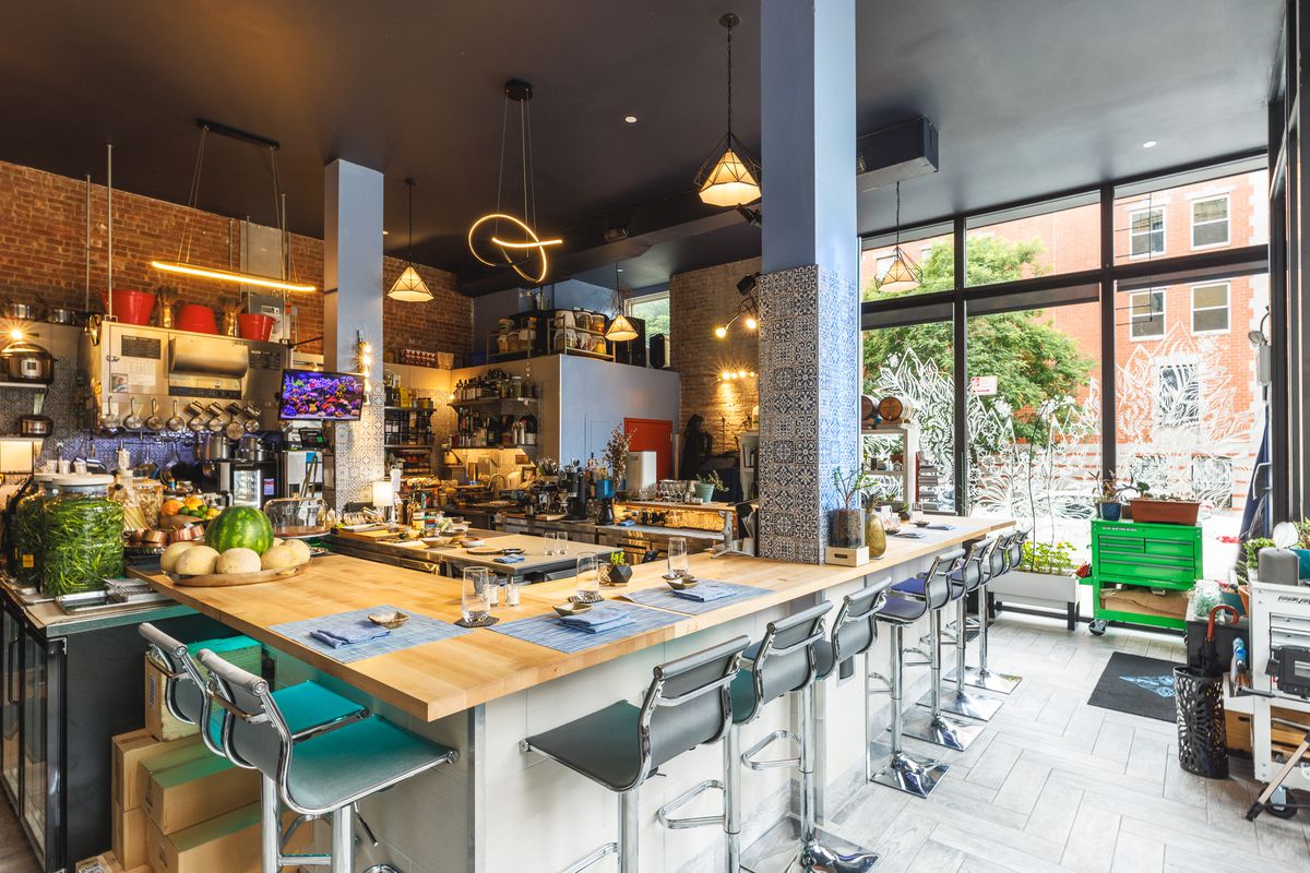 Upholstered steel bar stools with backs line the chef’s counter at Reverence before service; natural light floods in from the floor-to-ceiling windows