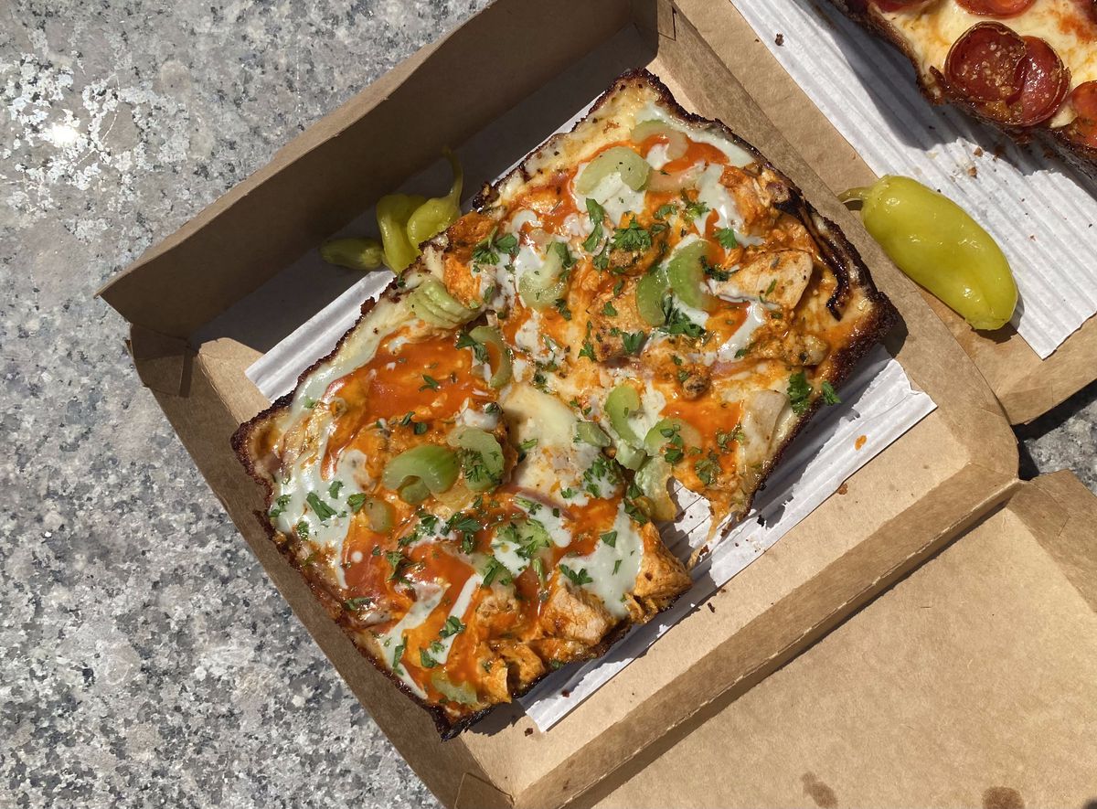 A slice of Detroit-style pizza covered in Buffalo sauce, chicken, and slices of celery sits in a cardboard box.