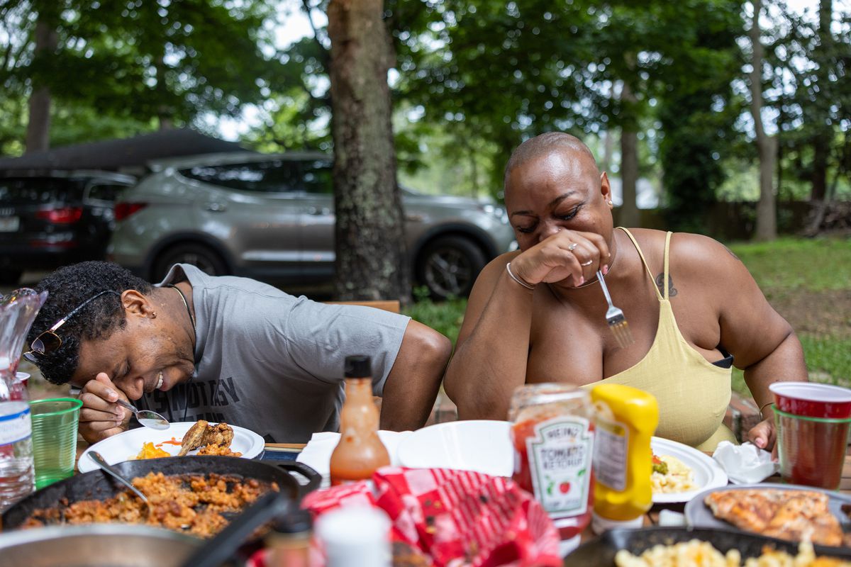 A man and woman crack up while sitting at a picnic table covered in food.