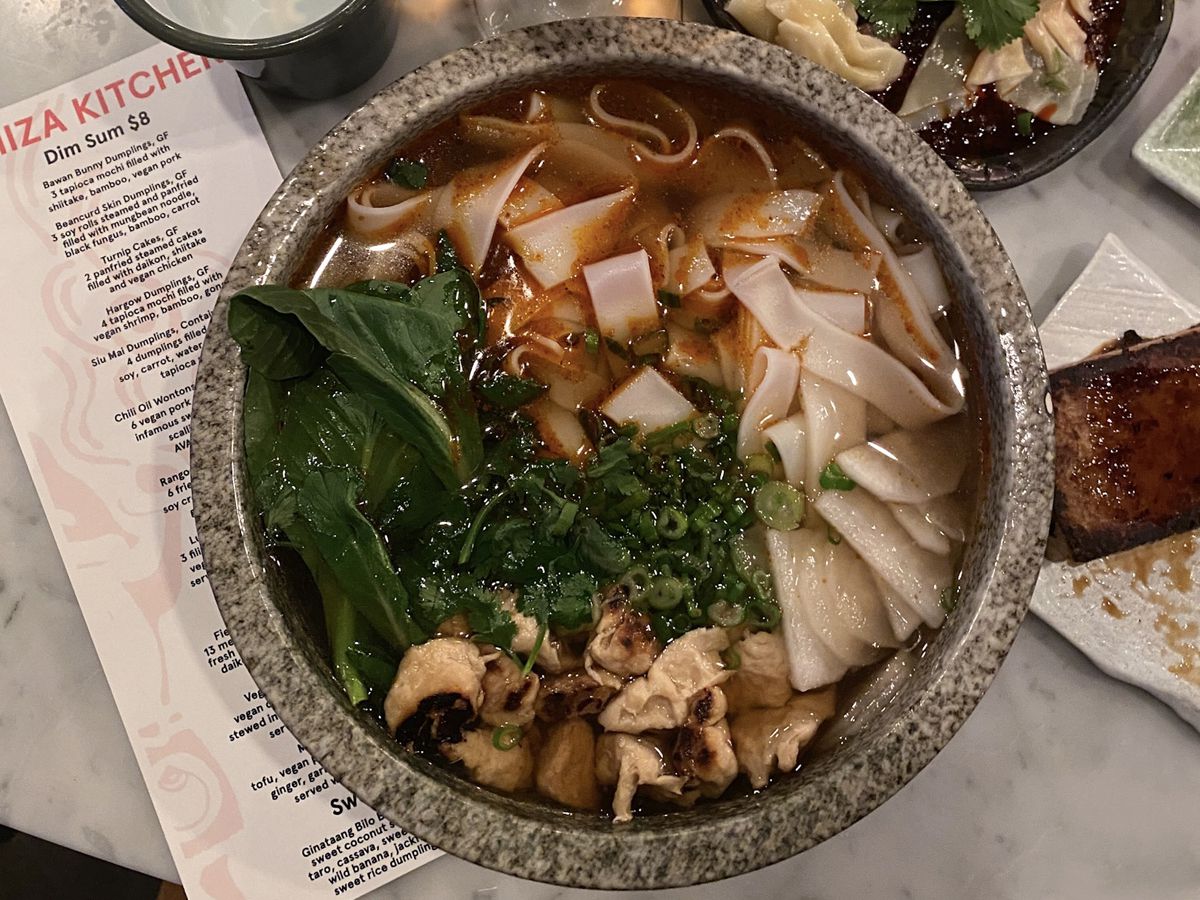 A photo of Jade Rabbit’s Fiery Wind noodle soup accompanied with turnip cakes, chile oil wontons, and a cocktail at The Emerald Room