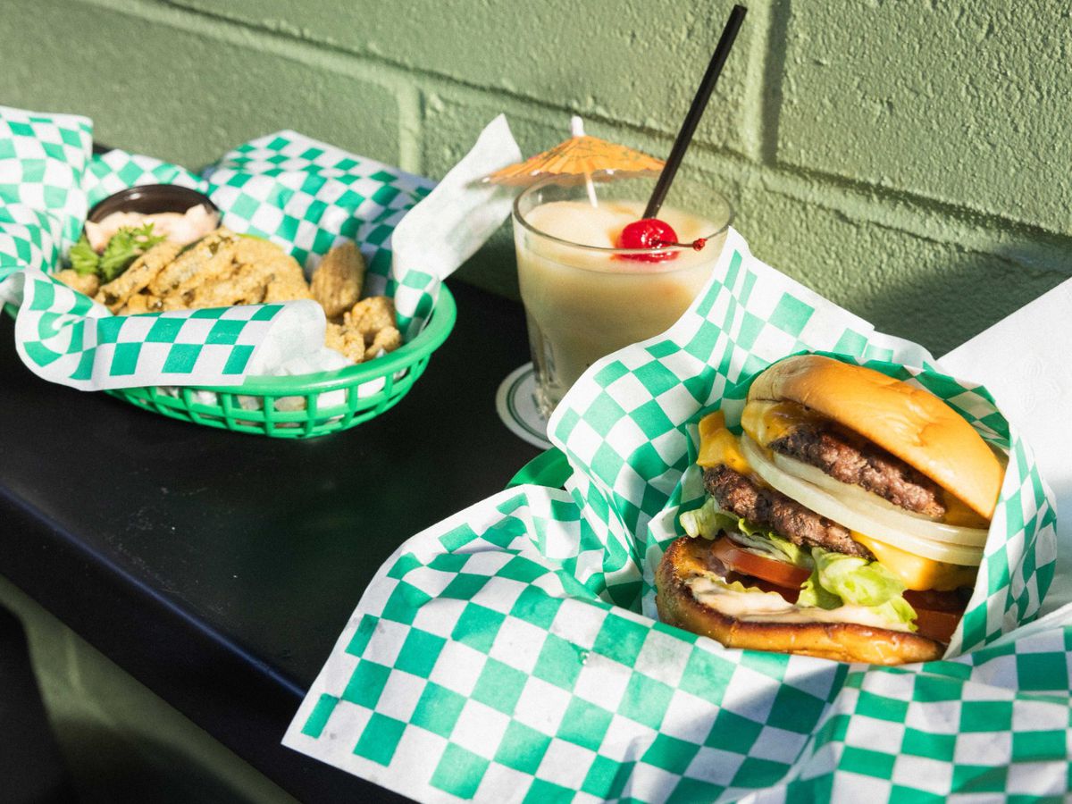 A basket of chicken tenders, a pina colada, and a double patty burger lined up on a black counter in green and white checkered paper.