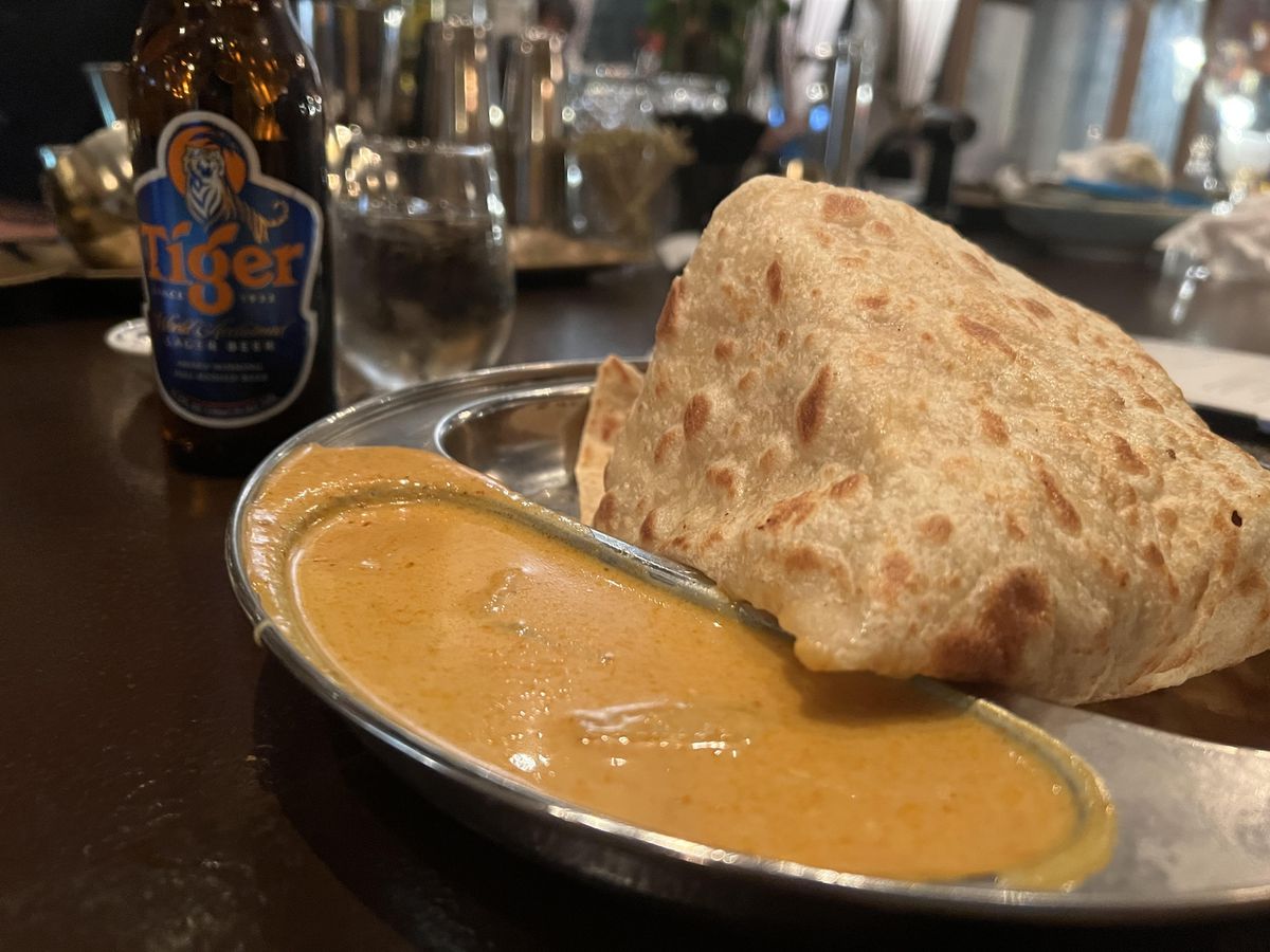 Golden roti canai flatbread sits in a metal tray next to a small pool of coconut curry