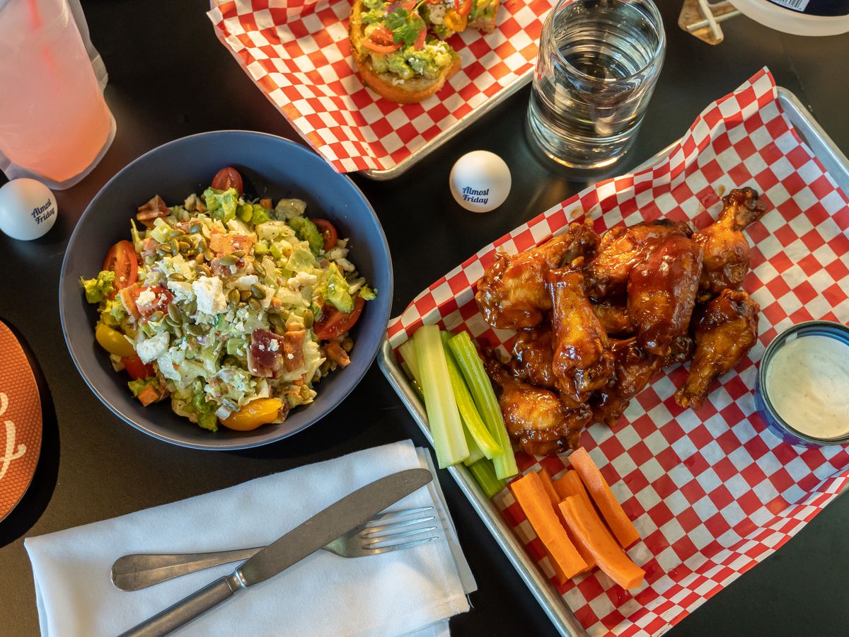 An overhead shot of a plate of chicken wings with celery and carrots on a gingham placemat, a salad bowl, drinks, and ping pong balls.
