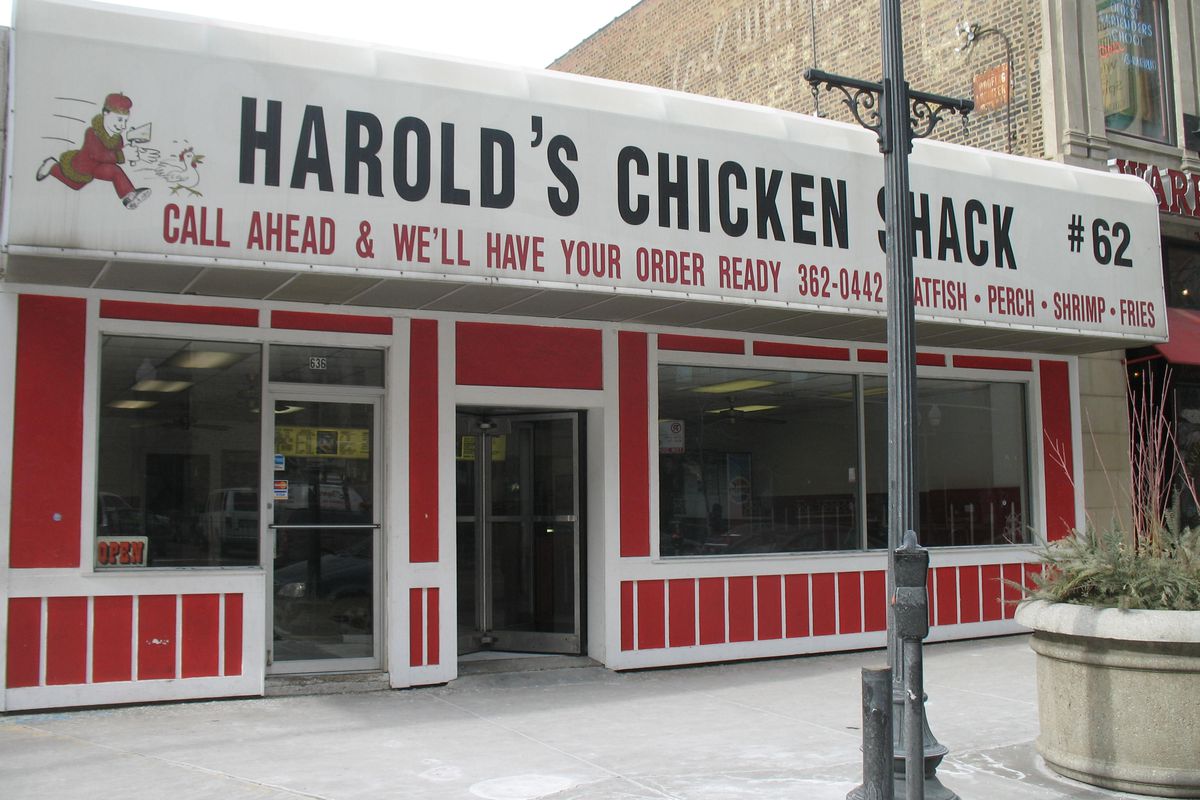 A restaurant with a red front and a white awning that says “Harold’s Chicken Shack #62” with the logo of a man chasing a chicken