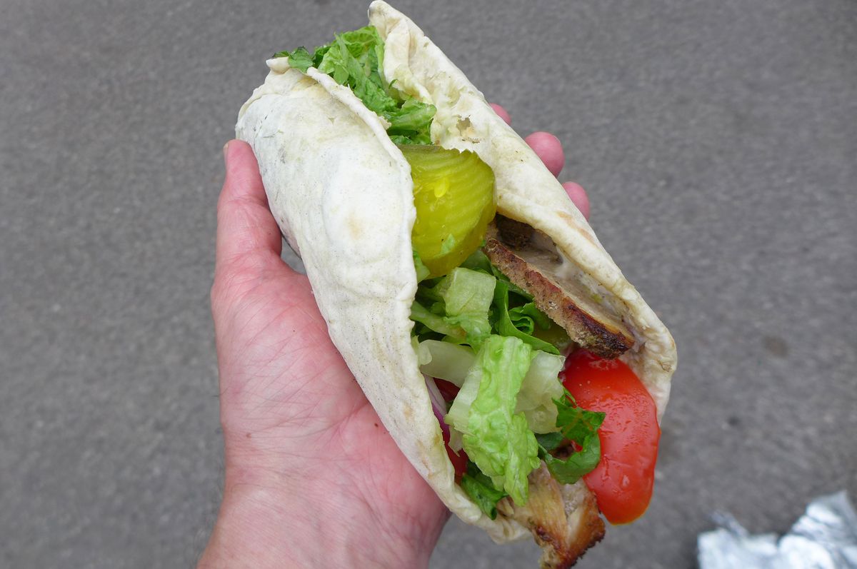 A hand holds a meat and lettuce sandwich with a pickle chip sticking out almost wrapped completely in a flatbread.