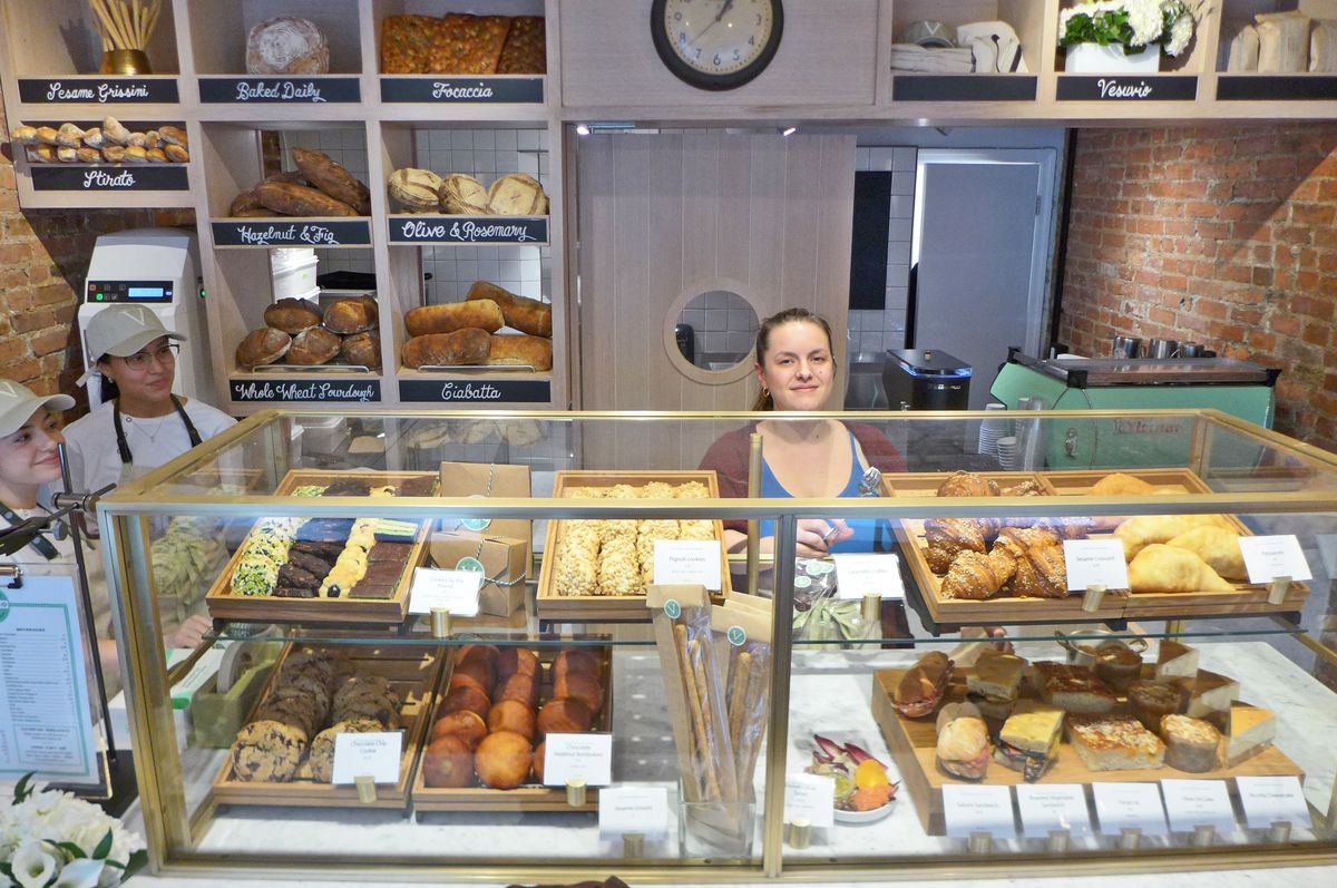 A woman stands behind a pastry counter as two clerks look on.