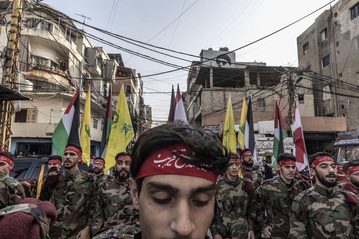 Young men, many bearded, all in fatigues and red headbands with white Arabic writing, march in a military formation, carrying Hezbollah and Lebanese flags. In the foreground is their leader, a clean shaven man, whose head is bowed and eyes closed. 