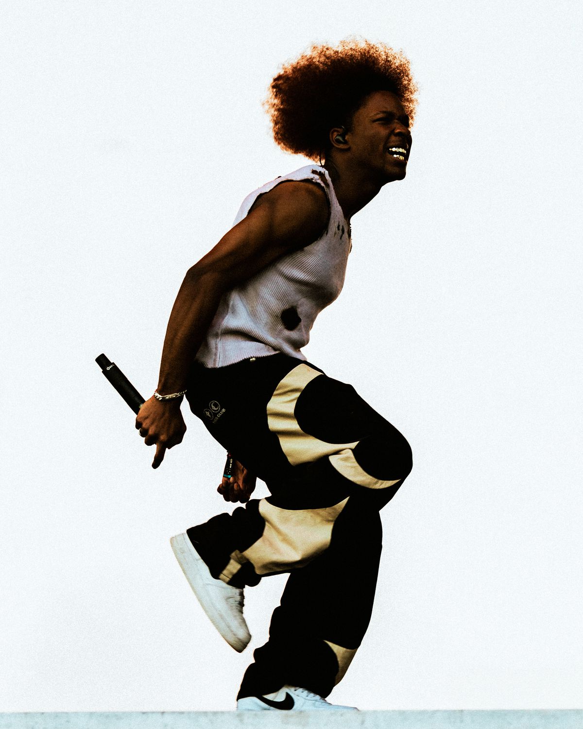 A person in a white tanktop and baggy black-and-gold pants holding a microphone. 