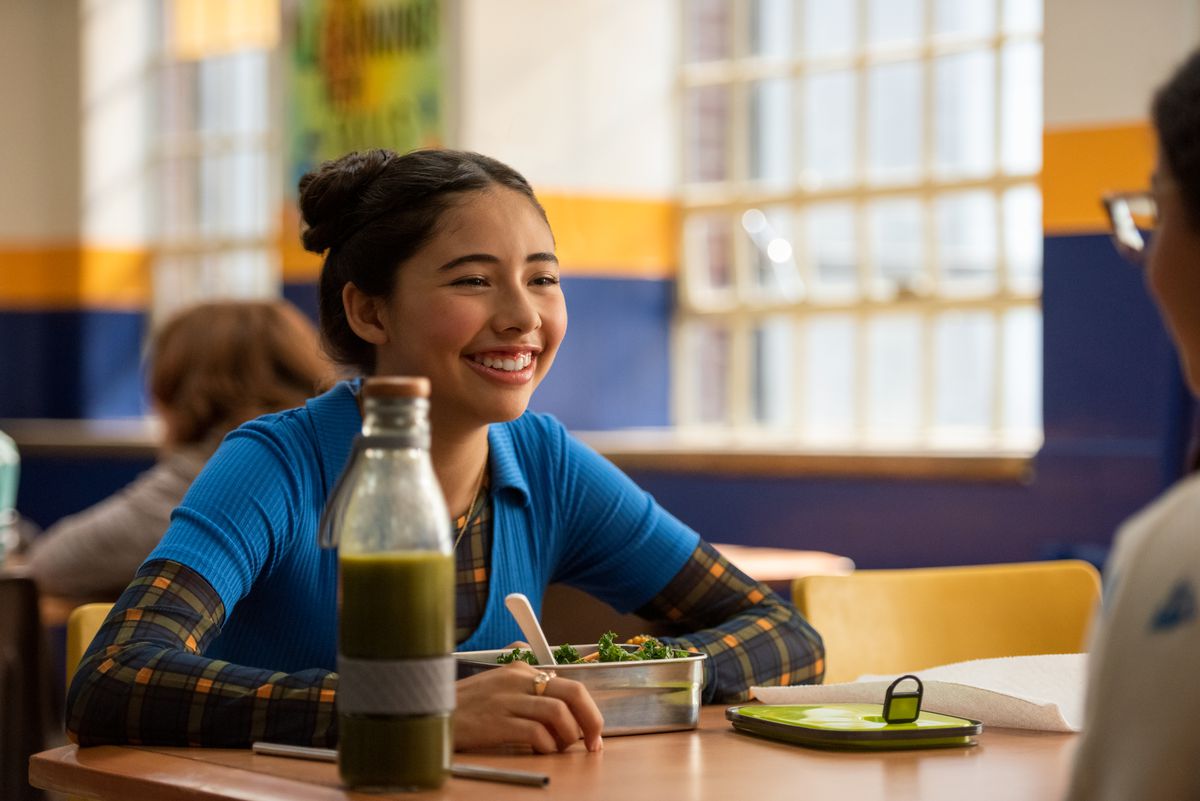 A Latina girl sits at a cafeteria table, smiling, with a salad and a bottle of green juice in front of her.
