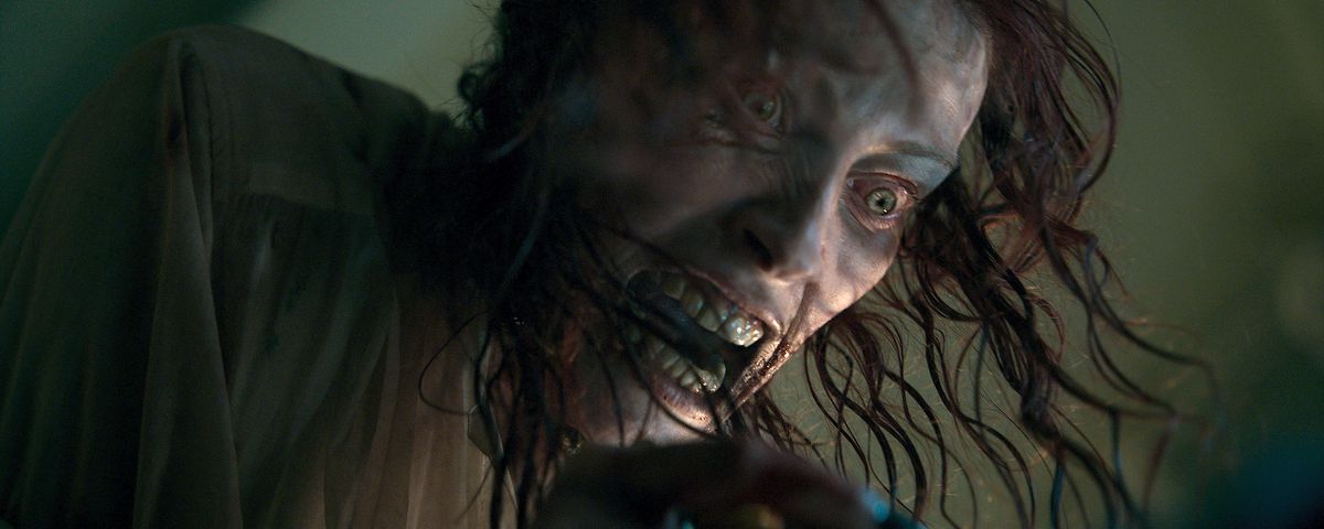Ellie (Alyssa Sutherland) dangles from the ceiling, stringy hair in her face, snarling and grey-skinned and grey-eyed, after being possessed in Evil Dead Rise