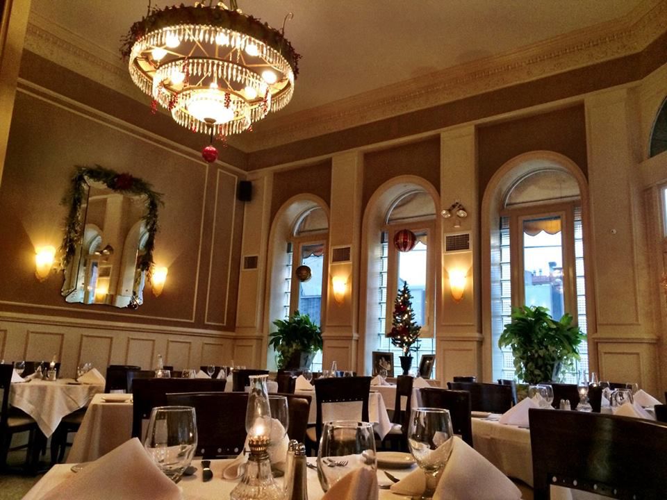The interior of a restaurant with white tablecloths, a chandelier, wall sconces, and tall windows.