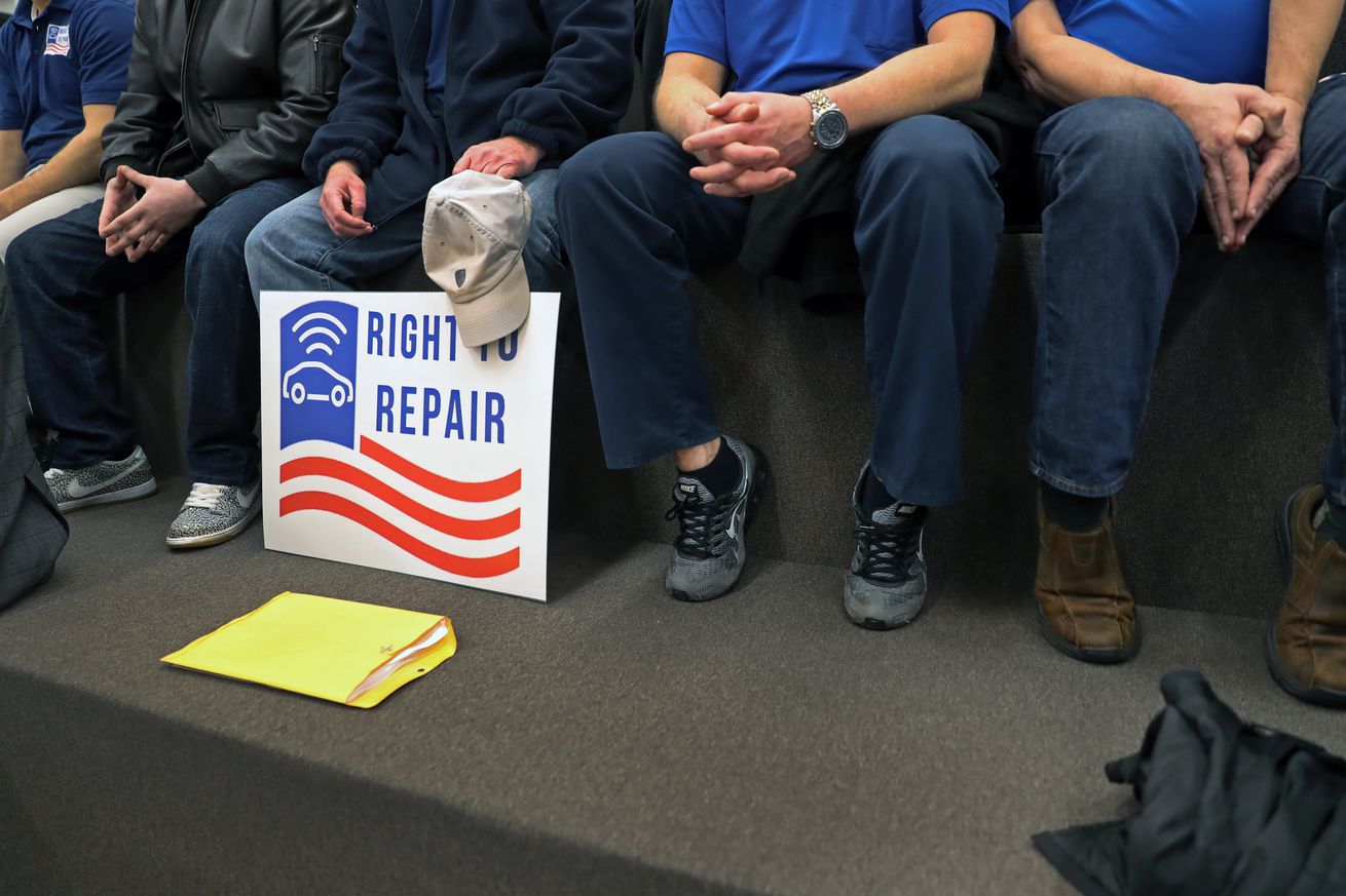 An image of several seated people from the waist down. A right to repair sign leans on a person’s knees.