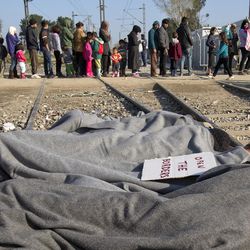 A banner is placed on a Syrian migrant sleeping on the railway tracks protesting the border closure and preventing freight trains from passing between Greece and Macedonia, as people wait for a daily ration of food, at the northern Greek border point of Idomeni, Greece, Sunday, March 20, 2016. German Chancellor Angela Merkel is urging migrants in the squalid tent city at Idomeni, on the Greek-Macedonian border, to trust Greek authorities and leave for better accommodation as thousands are still staying on site after the closure of Macedonia's border. 