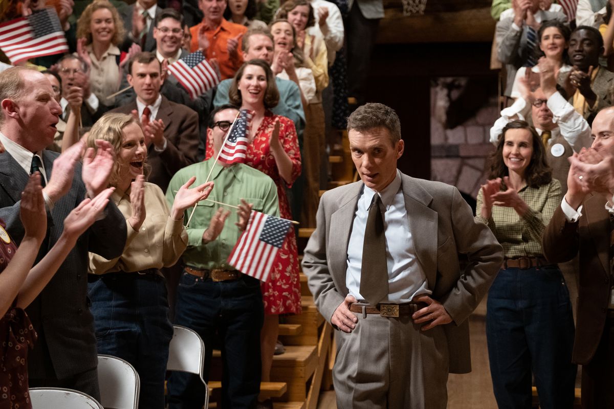 J. Robert Oppenheimer stands with his hands on his hips as he walks between twin bleachers filled with people cheering and waving American flags in a scene from the film Oppenheimer.