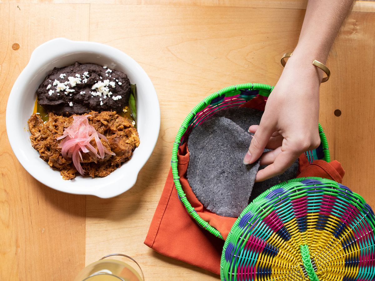 A white plate with brown shredded meat topped with pink pickled onions next to dark brown beans and person picking out a purple corn tortilla from a colorful open basket. 