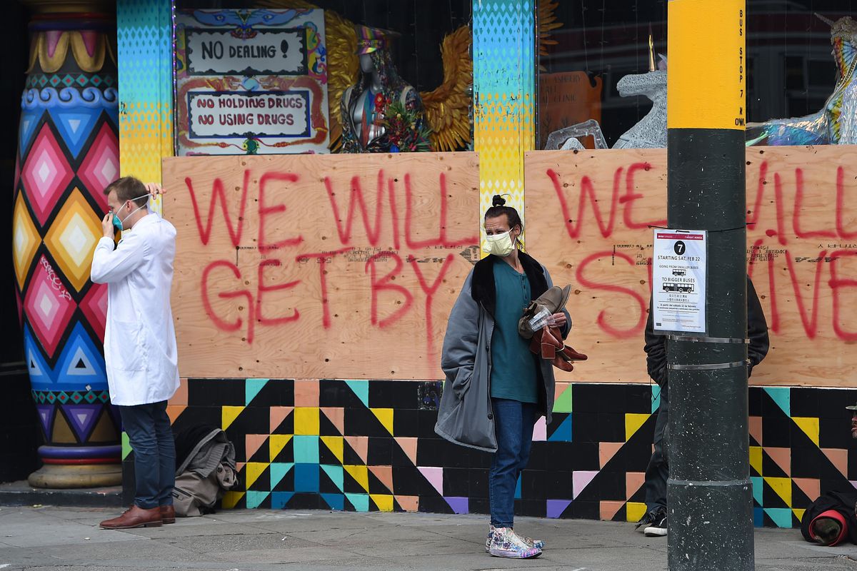 People in face masks stand in front of a hand-painted sign reading “We will get by. We will survive.”