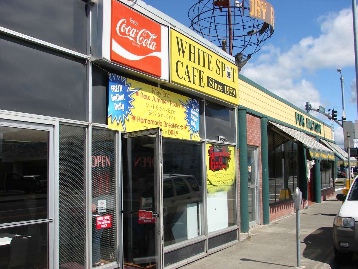 A row of building exteriors with dark windows. Above one door are bright signs, including one in yellow advertising Bright Spot Cafe, and a red Coca-Cola sign. 