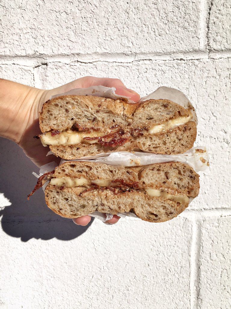 A hand holds a halved bagel sandwich in front of a white brick wall, showcasing the filling of almond butter, banana, honey, and bacon