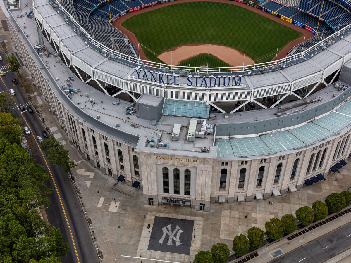An aerial view of the corner of a sports stadium. There is a baseball diamond within the stadium. On the outside of the stadium is the Yankees baseball team logo on the ground.