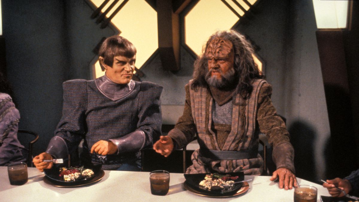 Costumed actors seated at a table with food and drinks in front of them.
