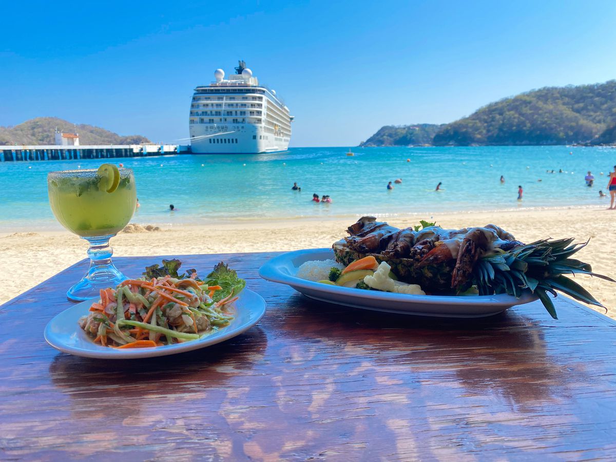 A table with several dishes, including a pineapple stuffed with seafood, and a cocktail overlooking a beach where a large cruise ships pulls in