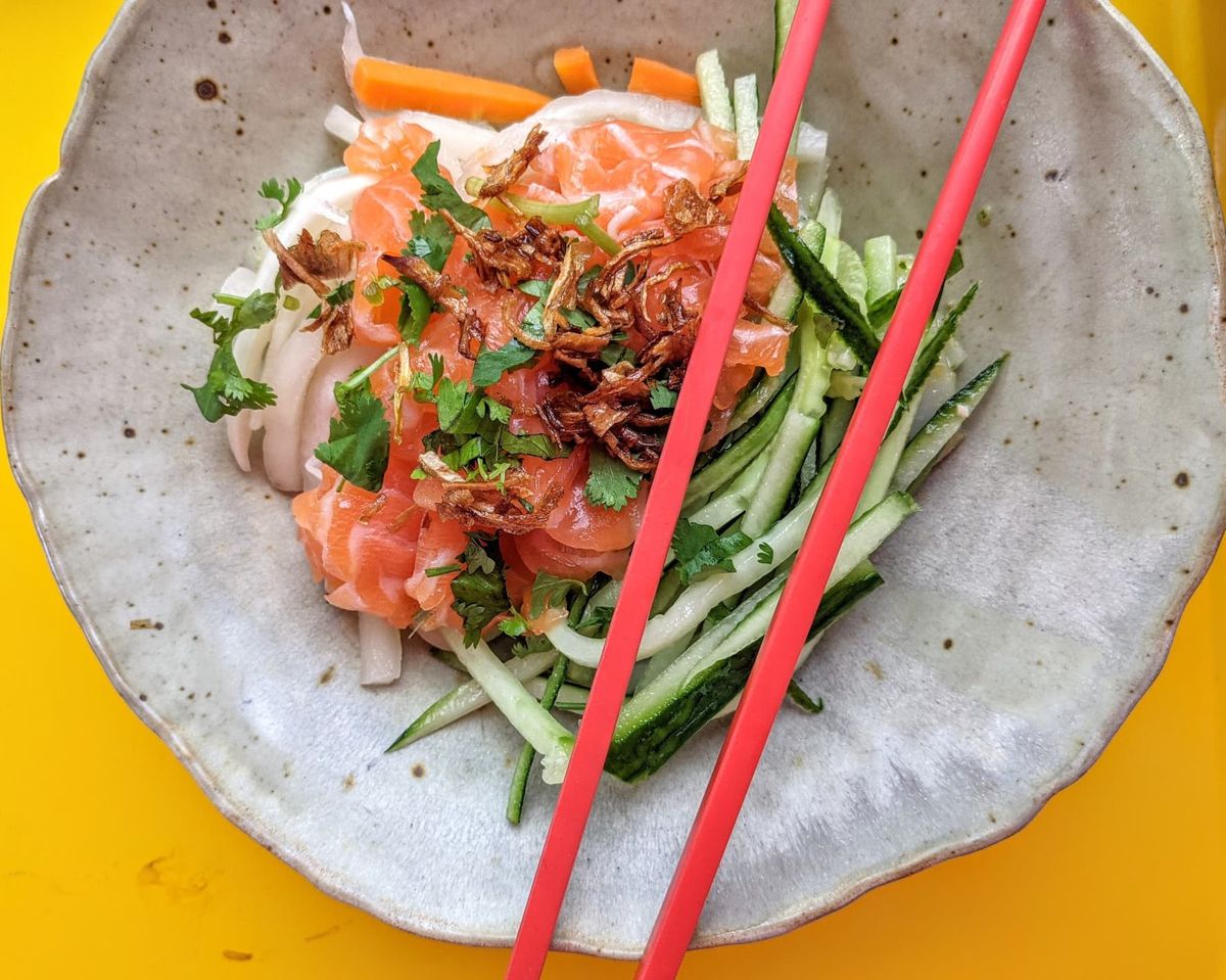 Overhead view of a ceramic bowl full of medium-thick rice noodles, long match-stick slices of cucumber and carrot, pieces of cured salmon, and a crispy topping. Two bright red chopsticks stretch across the bowl, which is on a bright yellow tabletop.