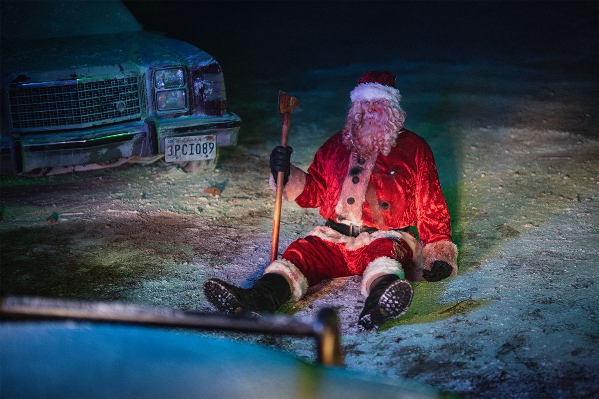 A man in a blood-covered Santa Claus outfit (Abraham Benrubi) holds an axe while sitting down in a snowy field next to a parked car.