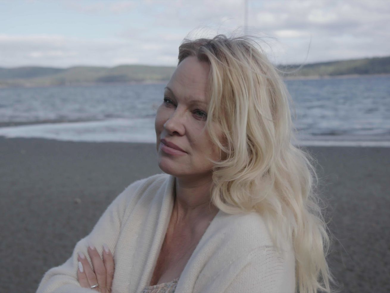 Actress Pamela Anderson stands on a beach, the ocean behind her. She is wearing a white sweater with her long blond hair loose, and her arms crossed, with a thoughtful look.  