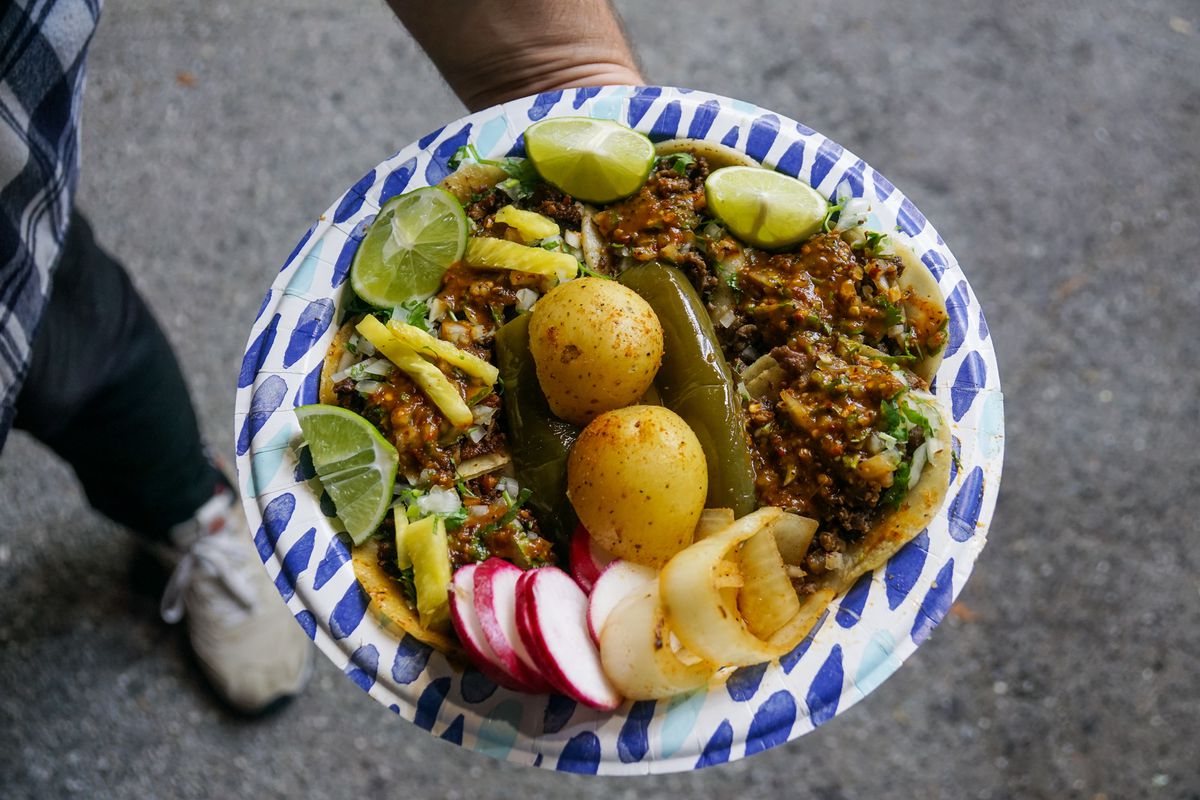 A hand holds up a sturdy paper plate ringed with tacos, limes, onions, grilled peppers, and potatoes in the center.
