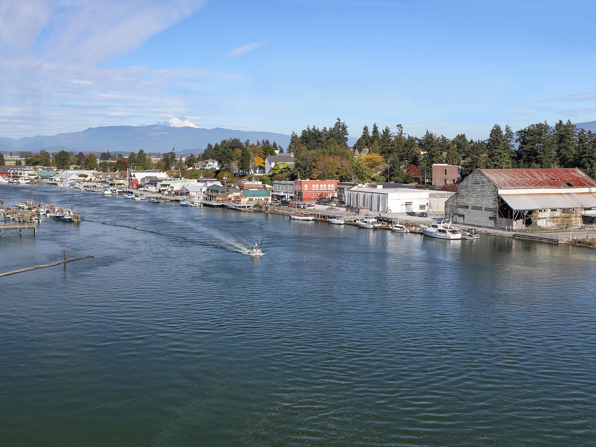 In the foreground is a body of water near Seattle. Lining the body of water are multiple houses and trees. In the distance are mountains. 