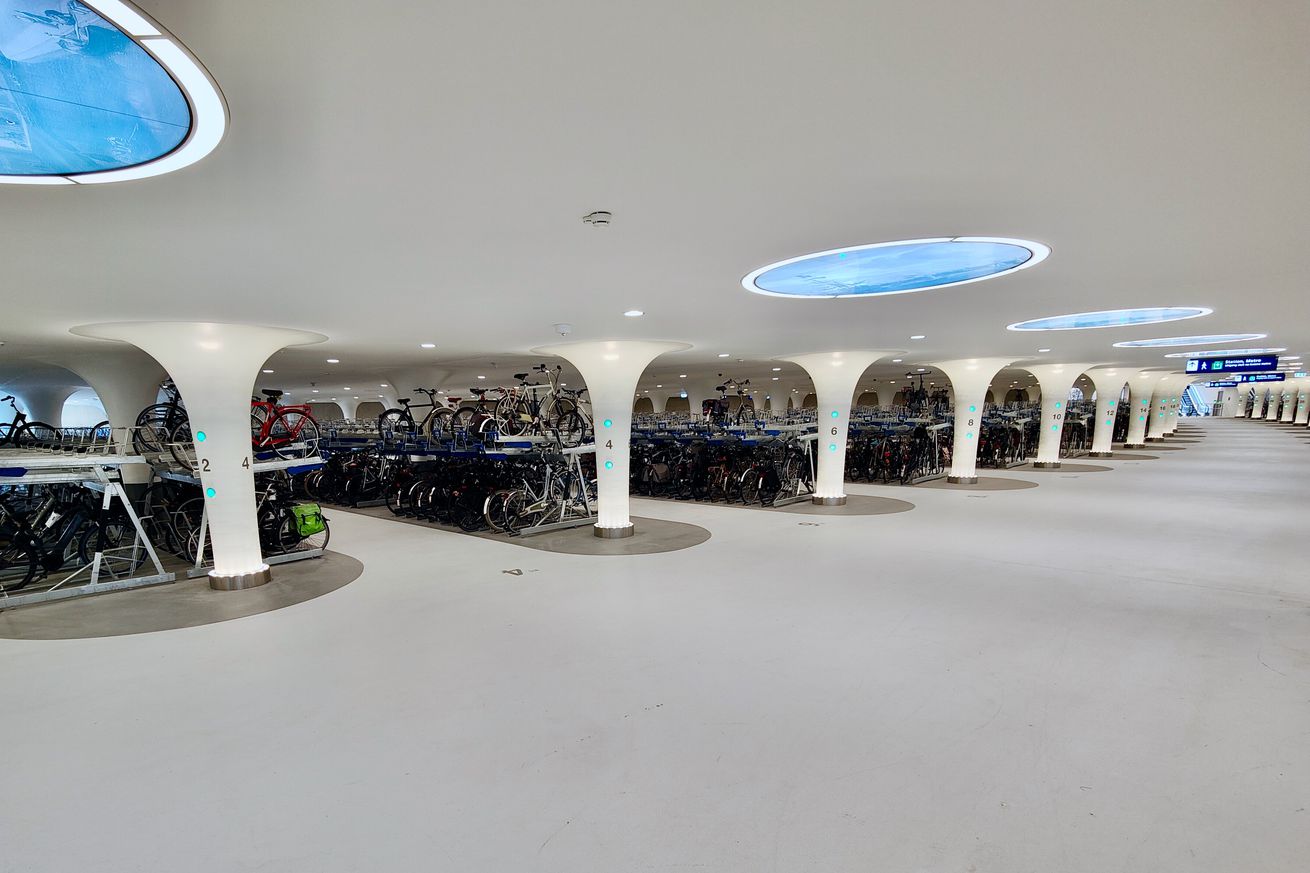 A photo showing a surprisingly spacious and futuristic parking garage of all white. Long rows of two-story bike racks extend to the edge of the room as tall white columns rise from the white floor to the white ceiling dotted with circular “windows” showing what looks like blue water (but are actually etched glass).