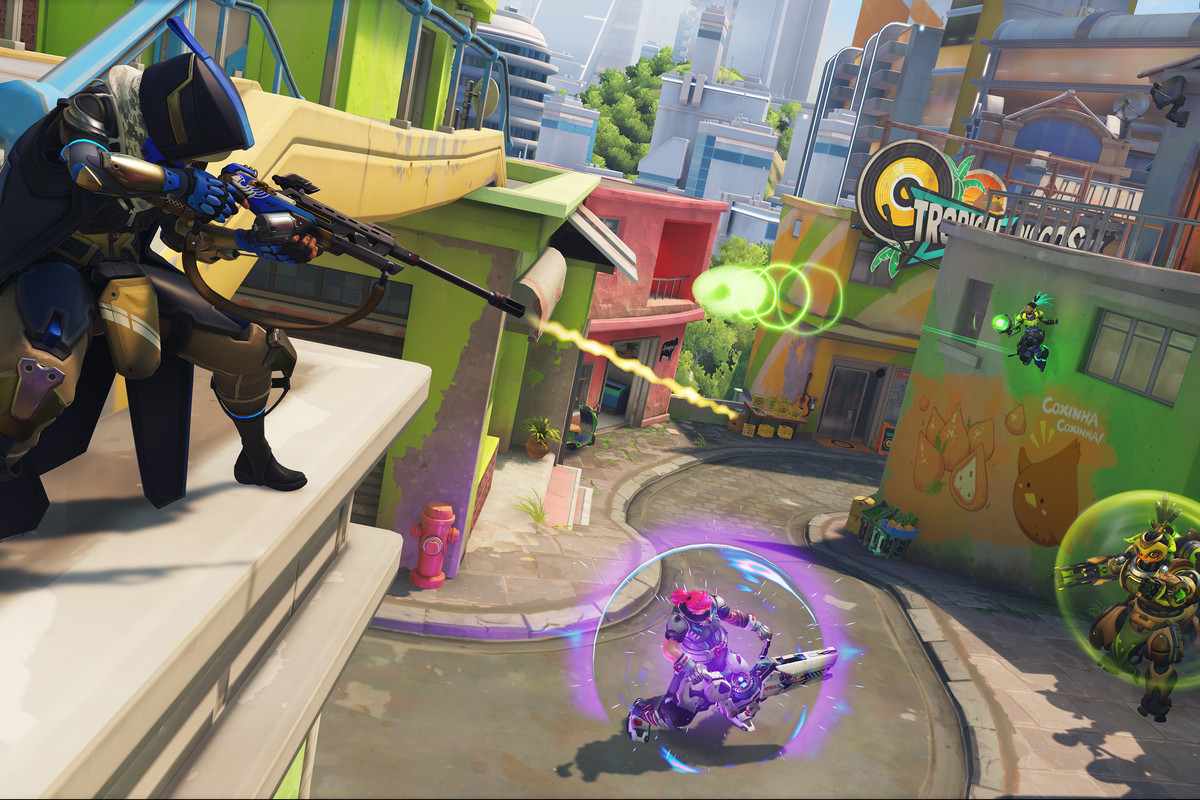 Ana, a sniper, stands on a rooftop and snipes down at a shielded Orisa robot. A variety of other heroes do battle on the colorful streets of Overwatch 2