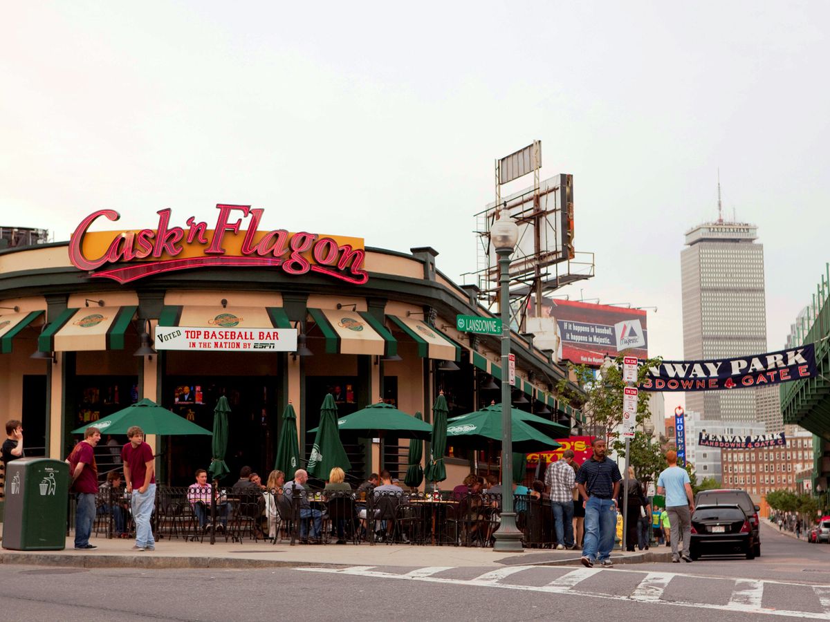 Exterior of a bar with green and beige accents and a large red sign reading Cask ‘n Flagon. A large banner for Fenway Park is stretched across the adjacent street.