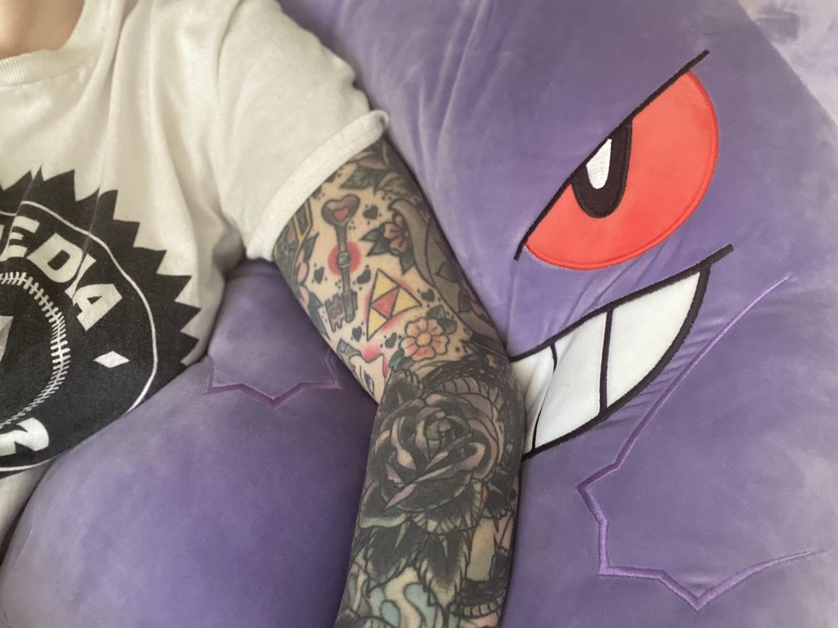 A woman laying on a Gengar Squishmallow, propped up by a tattooed arm.