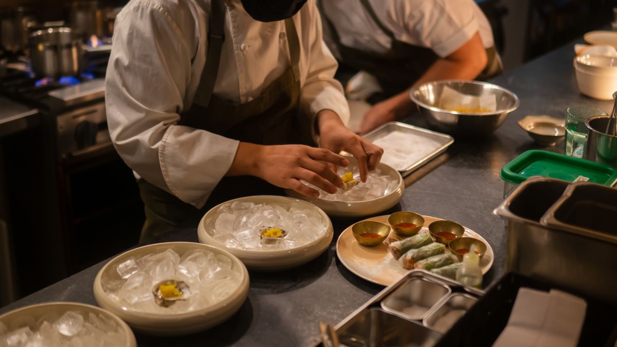 A chef wearing an apron and white shirt, and a black face mask, is plating three ice-filled shallow dishes on a kitchen.