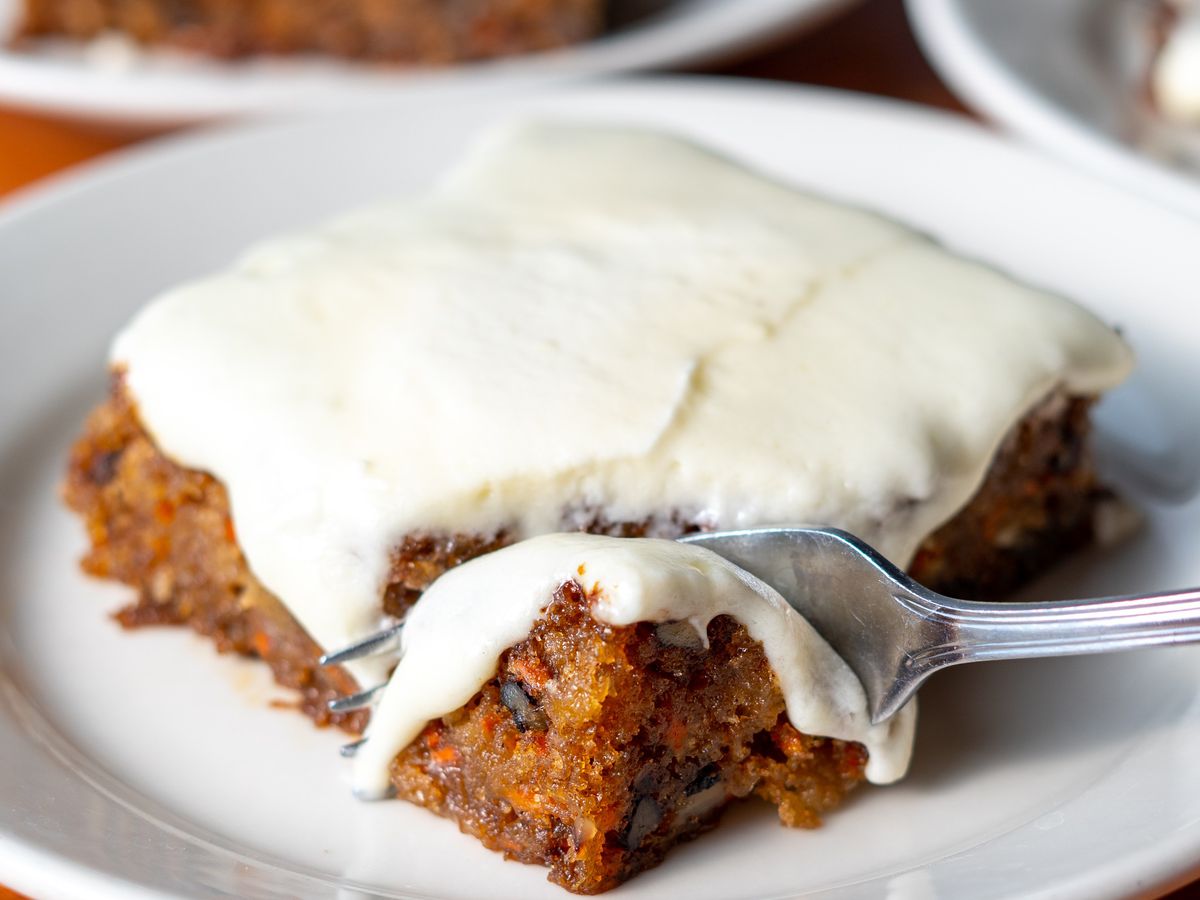 A fork scooping a bite of frosted carrot cake from a white plate