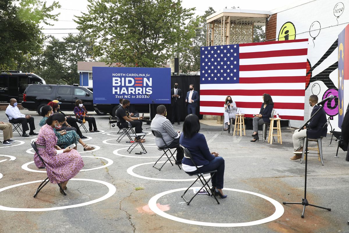 A group of people sit outdoors on black folding chairs; each chair is in the center of a white circle, and all the circle are six feet apart. Harris, in a grey suit and seated on a wooden stool, speaks with a woman in a black suit jacket and grey slacks; both of them are in front of a giant US flag. Next to the flag is a blue sign that says in white font, “North Carolina for Biden Harris Text NC to 30330.”