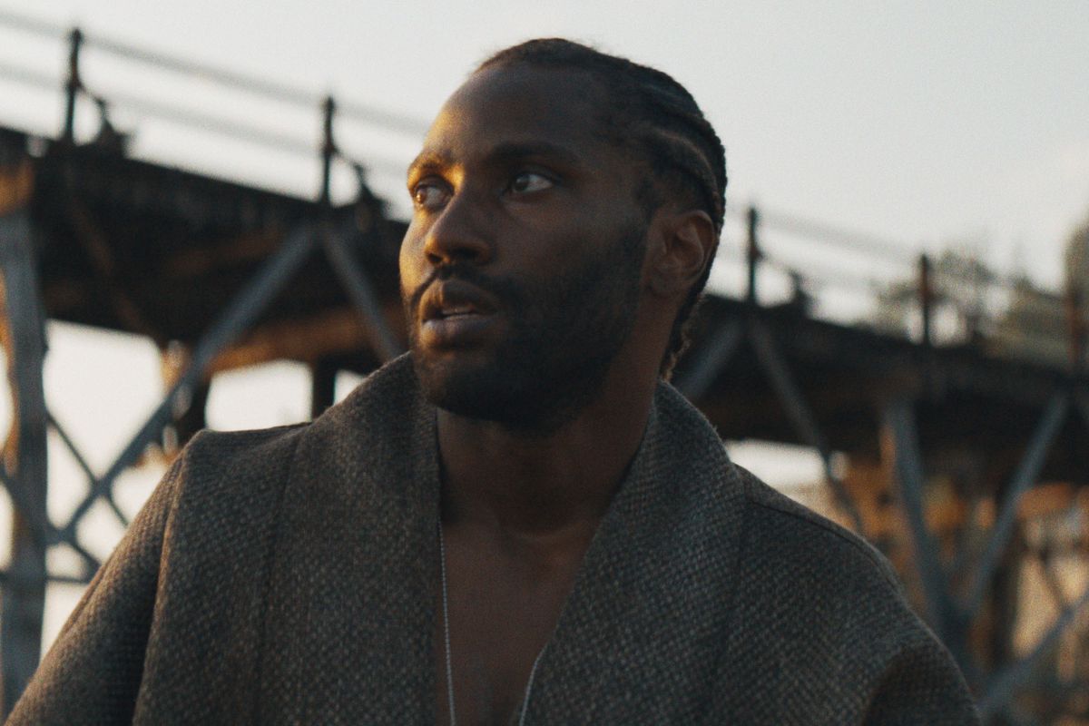 John David Washington as Joshua, looking up at something off camera as he stands in front of a wooden bridge at sunset in The Creator