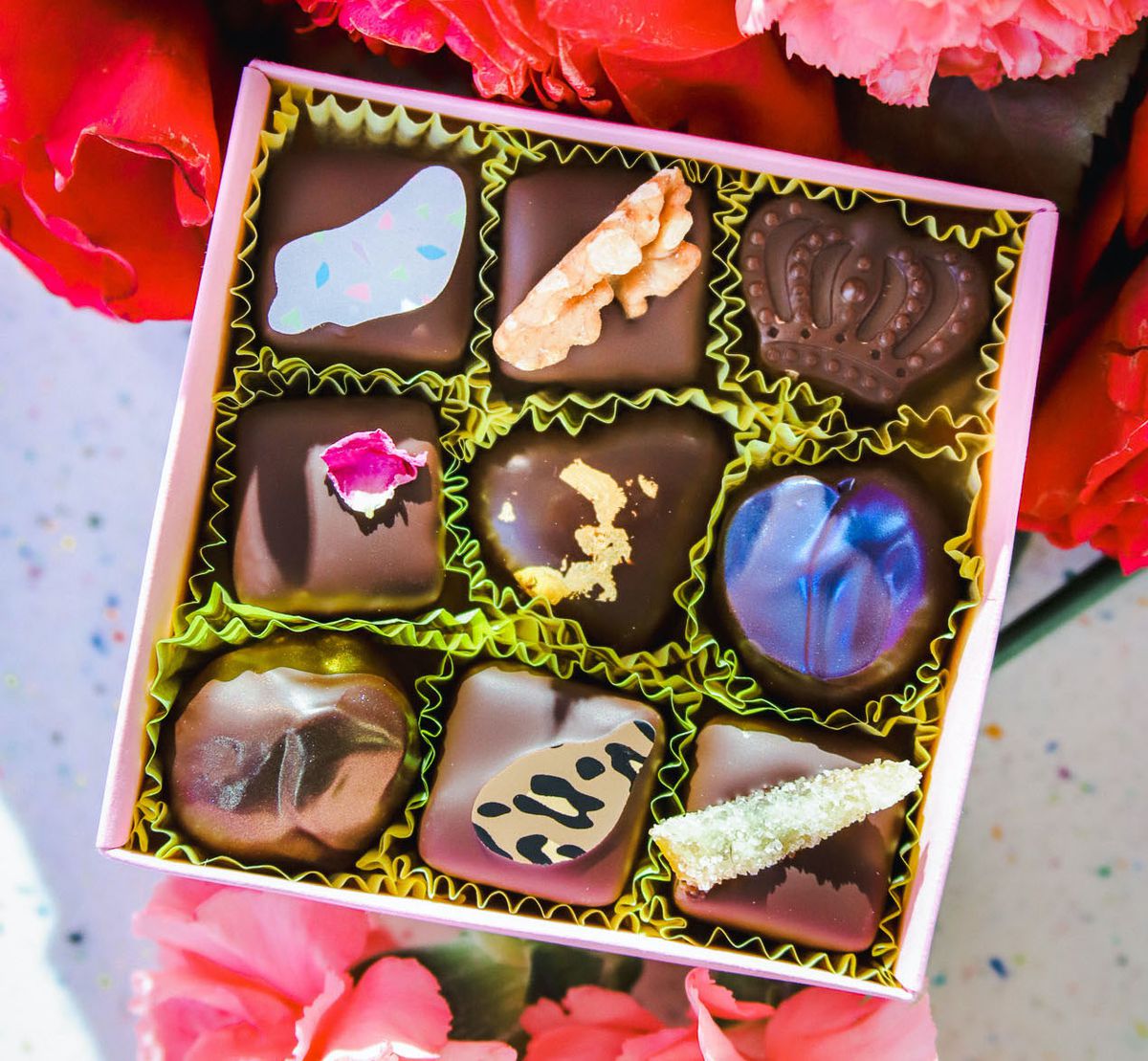 A small pink box is open to reveal nine fancy chocolate bonbons. It sits on a bouquet of roses.