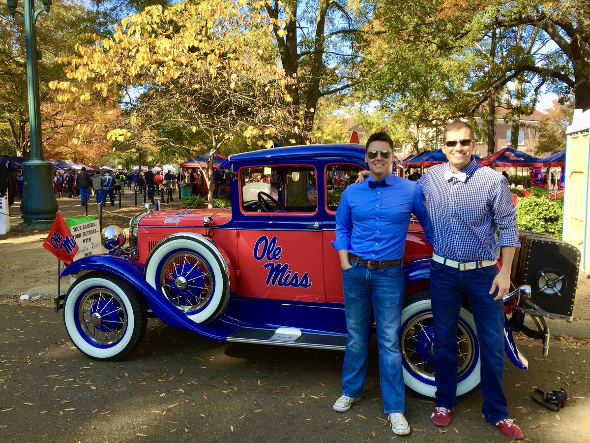 Married college football fans stand next to a classic car.