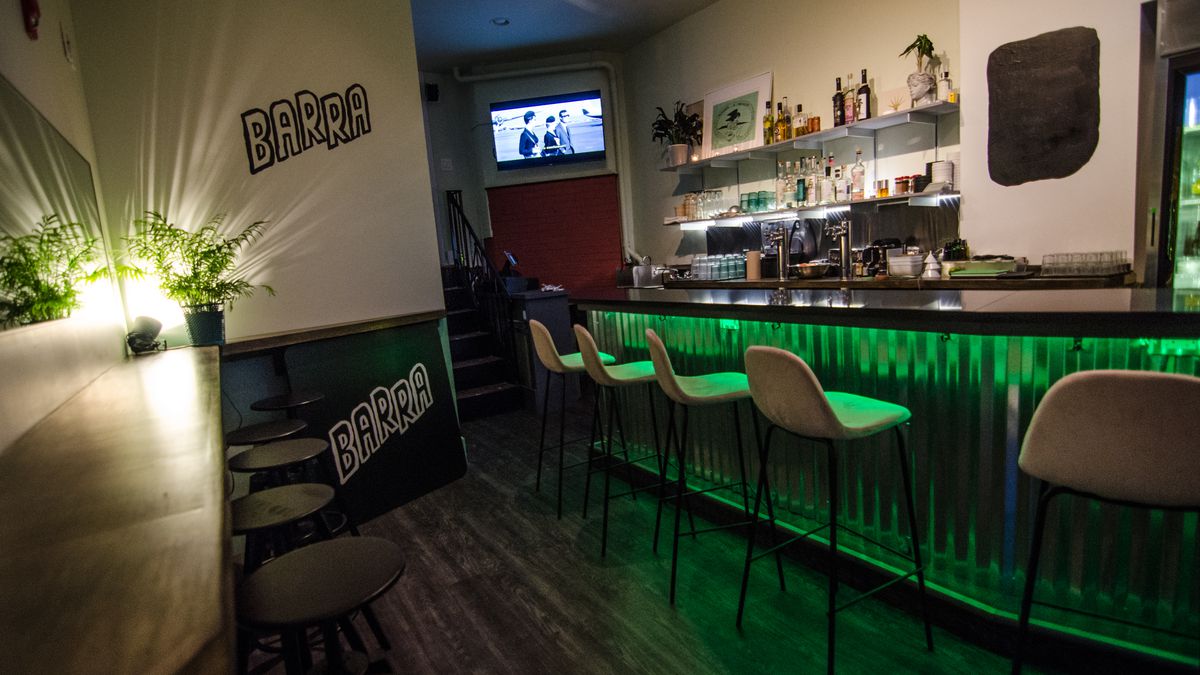 Interior of a small bar, dimly lit with green accents. The bar itself is lined with corrugated galvanized aluminum. The bar’s name, Barra, is hand-painted in thick black paint on the wall, and a spotlight casts lines through the leaves of a potted plant.