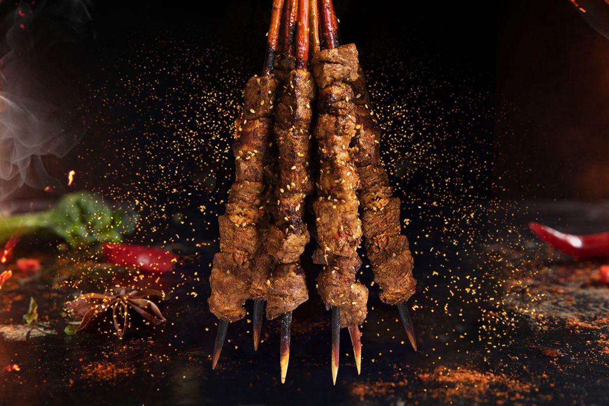 Six meaty skewers are held upside down, with peppers and spices coming off of them.