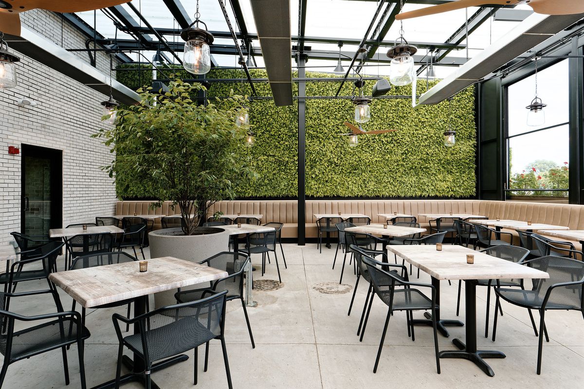 A patio with hanging lights and tables and chairs in the foreground and a wall covered with ivy in the back.