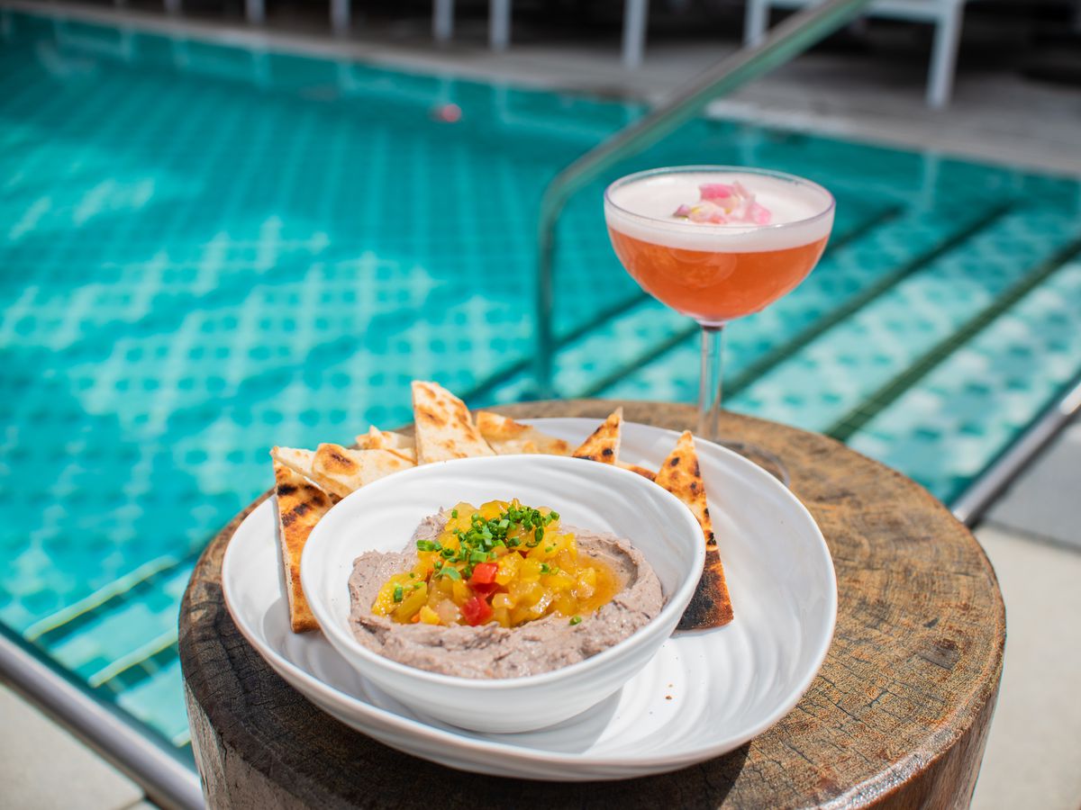 A white bowl of black bean hummus served with pita chips sits next to an orange cocktail in a coup glass on a wood stump table with a pool in the background.