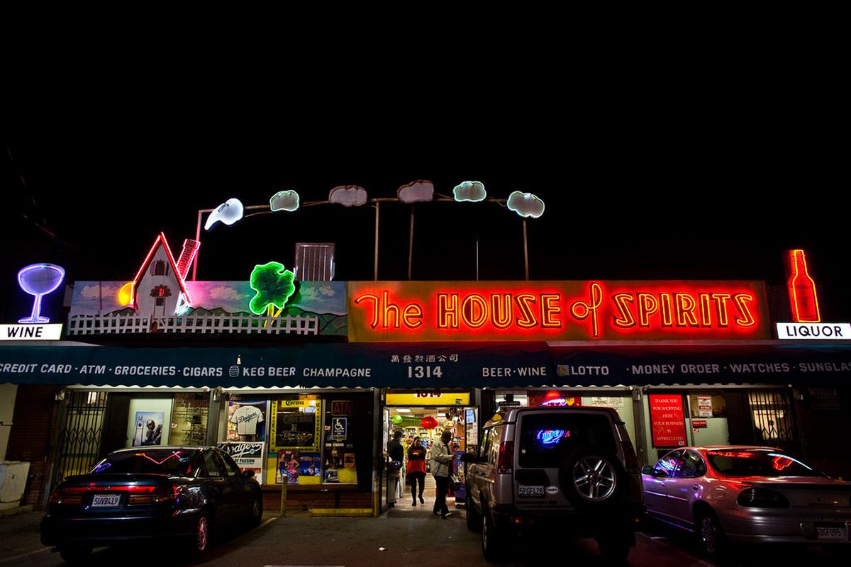 A nighttime photo of a liquor store with a green awning advertising available alcohol varieties (champagne, keg beer) and a neon sign that advertises the store’s name shows a house with a chimney with puffs of smoke coming out.