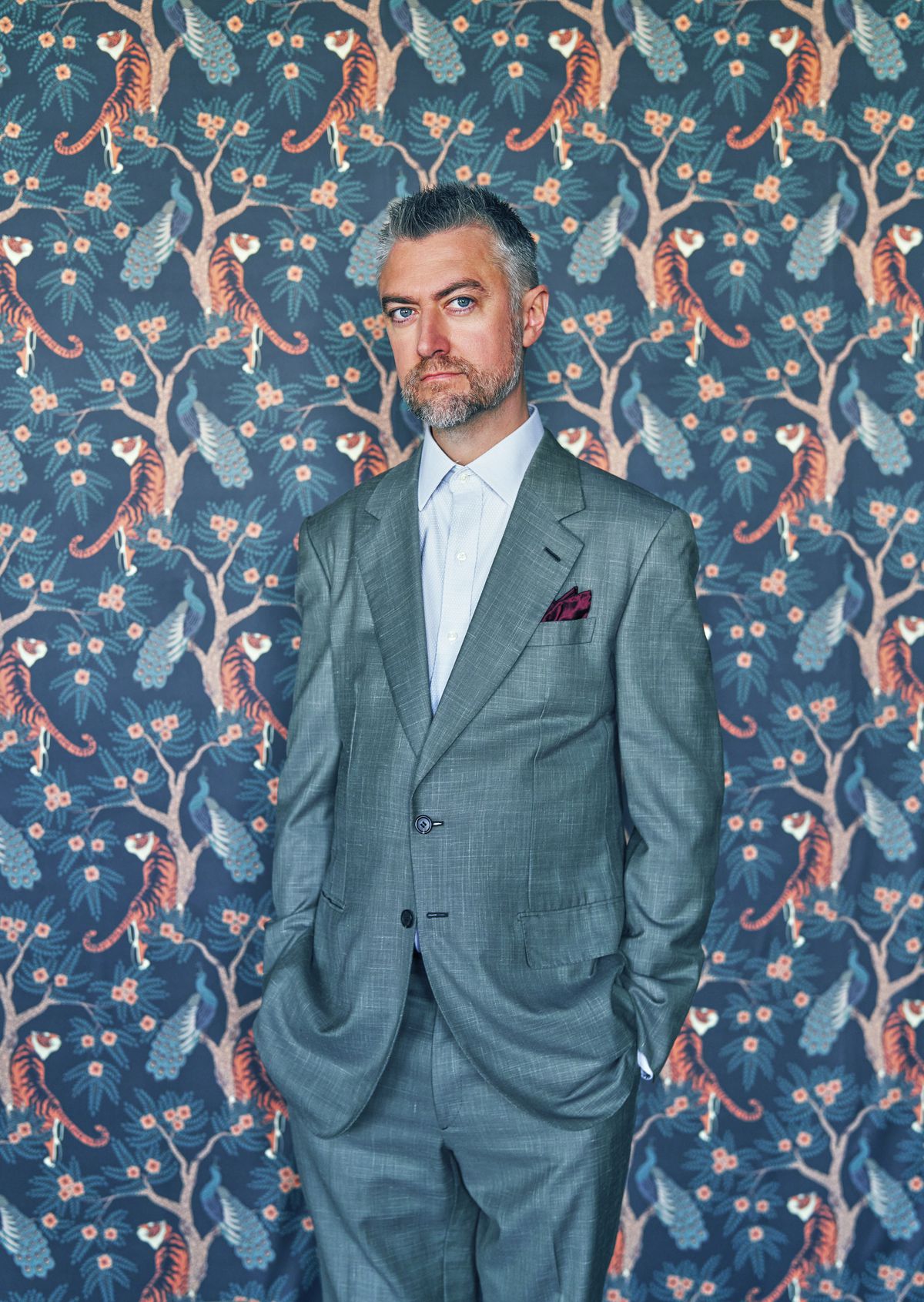 Actor Sean Gunn in a blue suit, standing against a busy background with a repeated pattern of peacocks in trees with tigers below