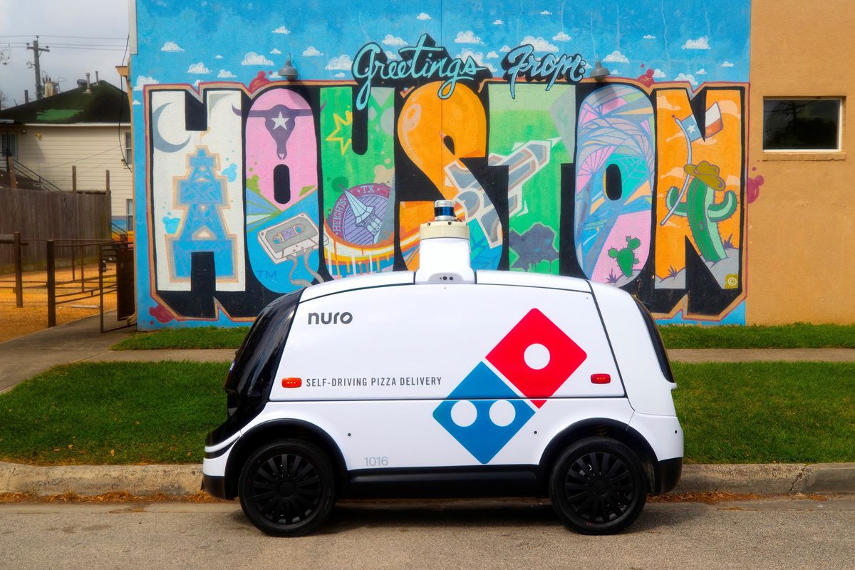 A van-resembling autonomous vehicle with a Domino’s logo painted on its side, in front of a Houston mural.