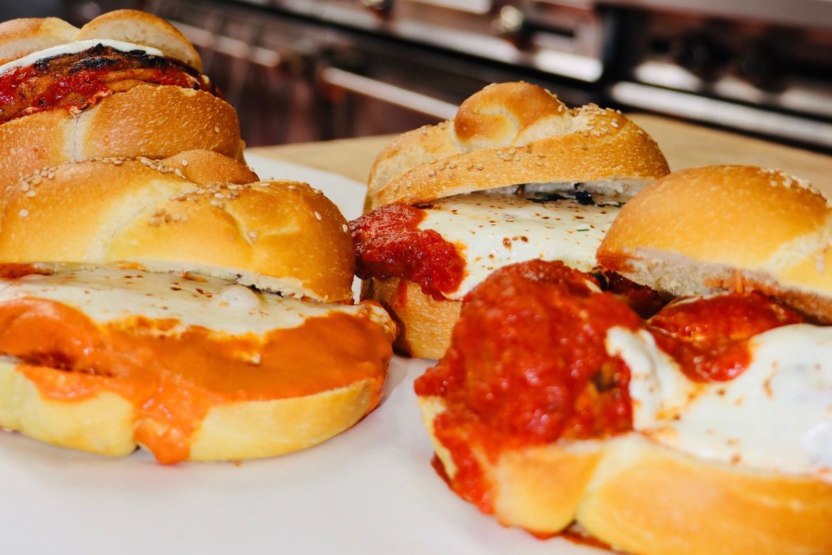 Four parm roll sandwiches with melty cheese and tomato sauce sit on a tray. In the background, an industrial kitchen oven is visible.